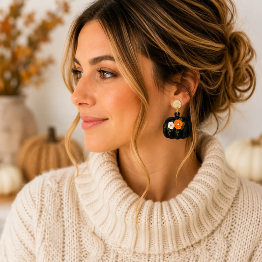Model wearing lightweight black pumpkin dangle earrings featuring orange and white floral details, green vines, and gold-tone studs.