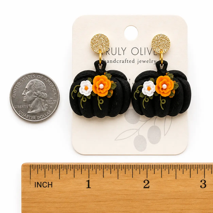 Size comparison of black pumpkin dangle earrings with orange and white floral accents and green vines. It shows the earrings size compared to a quarter. Showing the approximate length and width of the pumpkin earrings