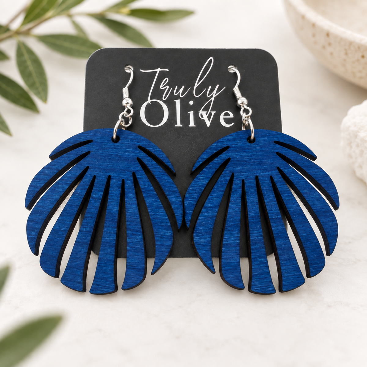 Blue wood leaf earrings on a Truly Olive card, styled on cream linen with softly blurred greenery.