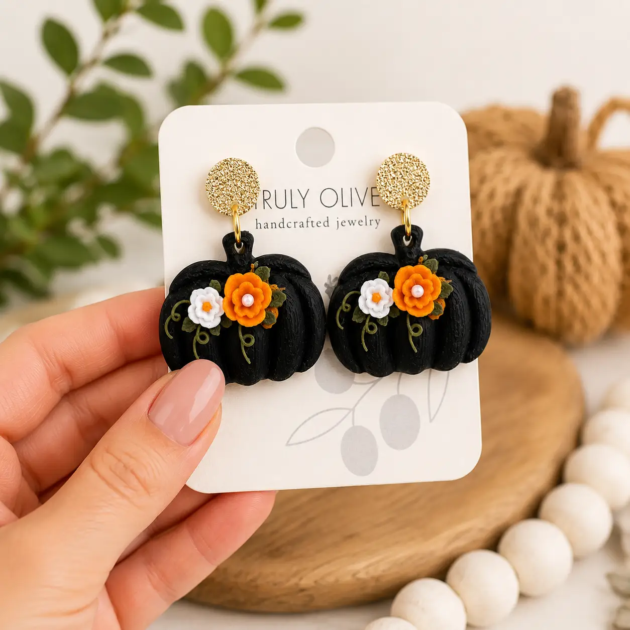Hand holding black pumpkin statement earrings with layered orange and white flowers, green leaf accents, and textured gold stud tops.
