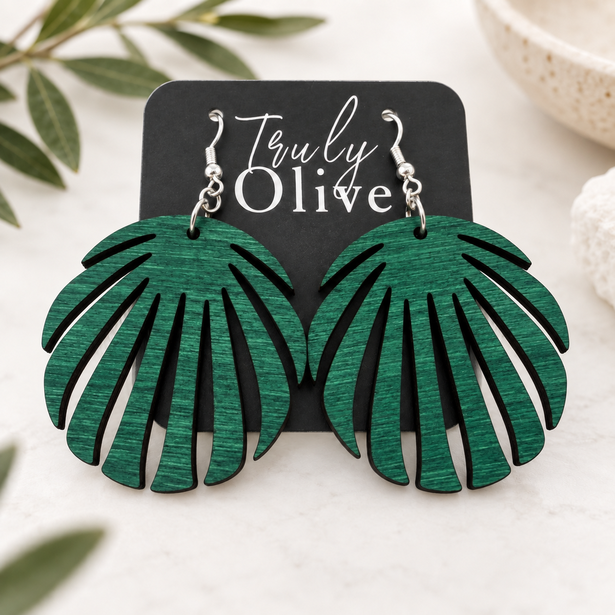 Green wood leaf earrings on a Truly Olive card, styled on cream linen with softly blurred greenery.