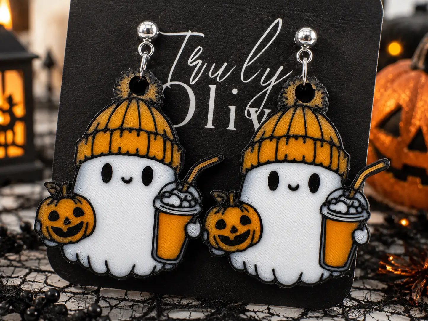Cozy ghost Halloween earrings displayed on a Truly Olive jewelry card surrounded by pumpkins, warm lights, spiderweb décor, and festive orange-and-black Halloween accents.