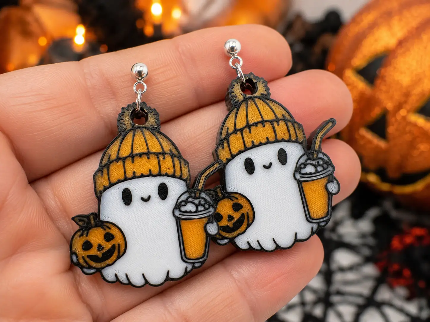 Pair of handcrafted ghost earrings resting in an open palm, showing white ghosts, orange knit hats, smiling pumpkins, fall drinks, and silver-tone stud hardware.