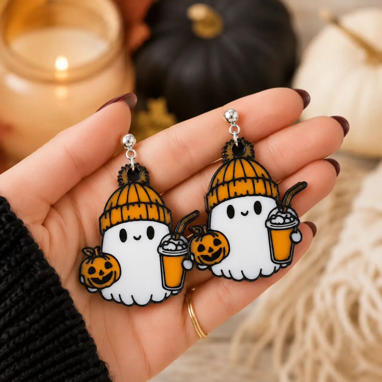 Cute little handcrafted ghost earrings resting in an open palm, showing white ghosts, orange knit hats,  holding a mini smiling pumpkins, fall  latte coffee drink, and silver-tone stud hardware.