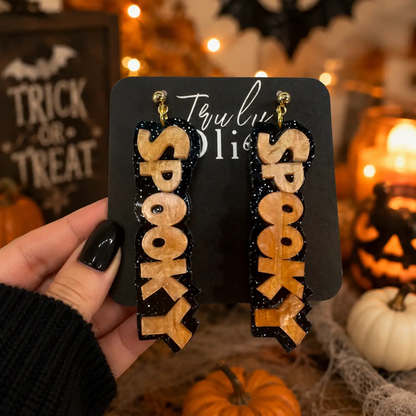 Hand holding orange and purple SPOOKY statement earrings on Truly Olive cards with Halloween decorations in the background.