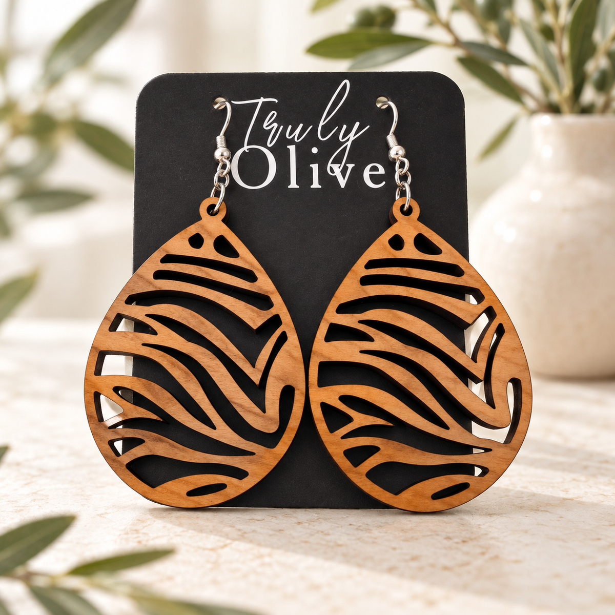 Cherry wood earrings on a black Truly Olive card atop light stone, with softly blurred olive greenery and a cream vase.