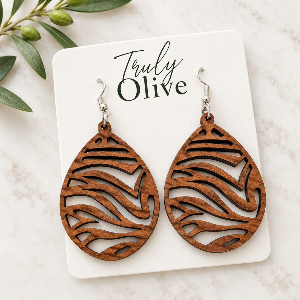 Overhead view of Sapele wood earrings on an angled white Truly Olive card beside an olive sprig on light stone.