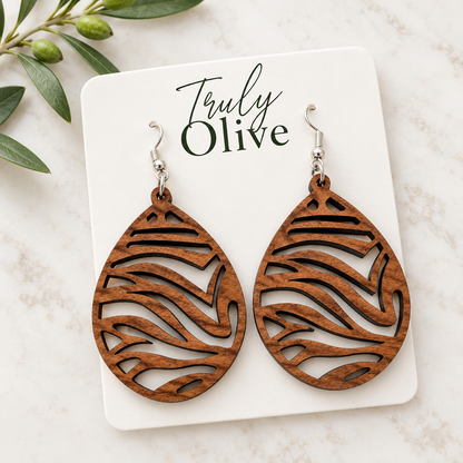 Overhead view of Sapele wood earrings on an angled white Truly Olive card beside an olive sprig on light stone.