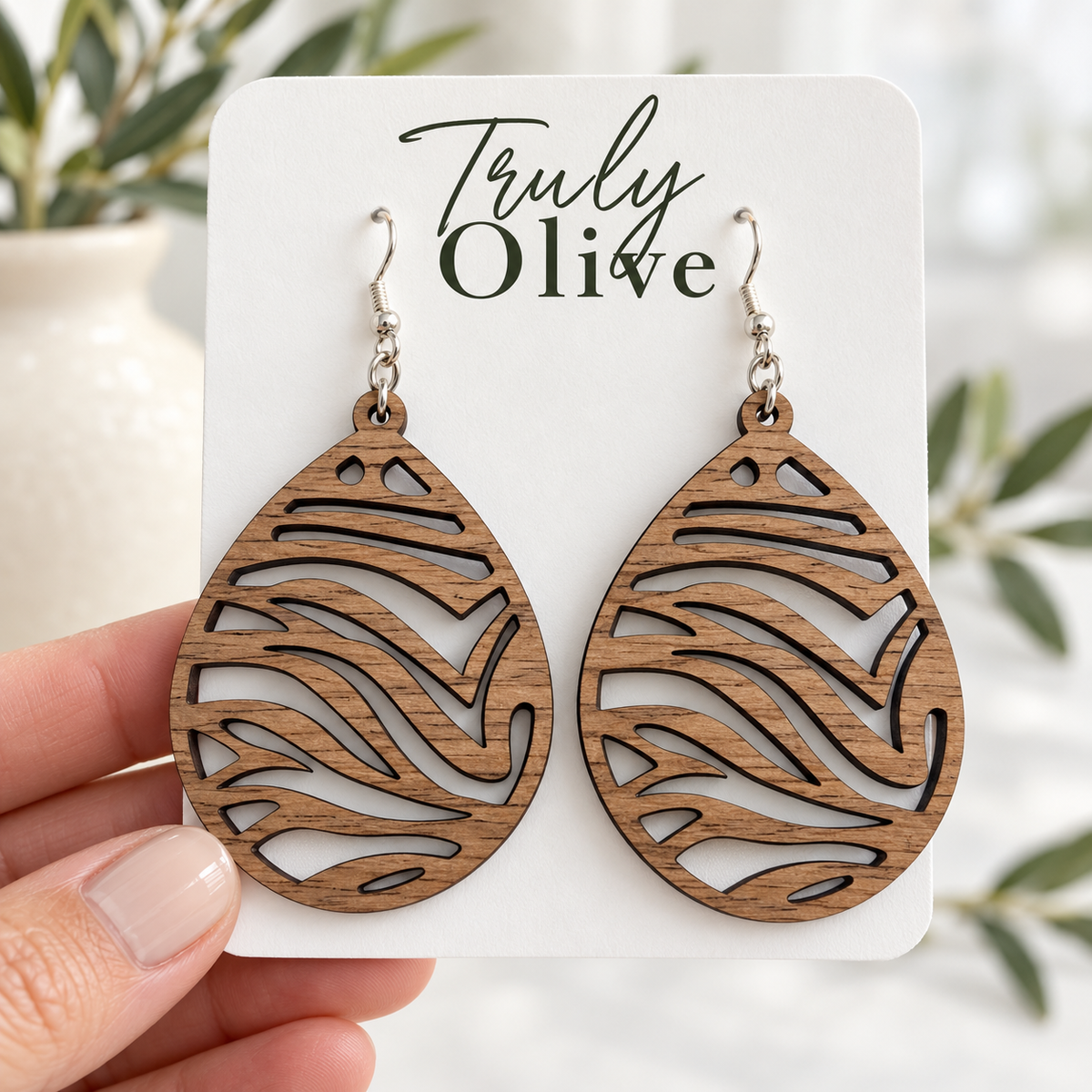 Hand holding a white Truly Olive card displaying Walnut wood zebra cutout earrings against a soft boutique background.