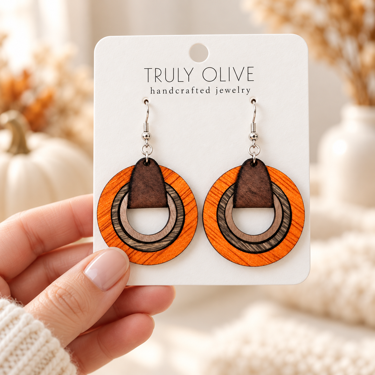 Hand holding a white Truly Olive card displaying orange and neutral brown wooden hoop earrings.